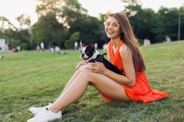Mutlu güzel bir kadın yaz parkında çimenlerde oturuyor, elinde Boston Terrier köpeği, gülümsüyor pozitif ruh hali, turuncu elbise giyiyor, moda tarzı, ince bacaklar, spor ayakkabıları, evcil hayvanlarla oynuyor.