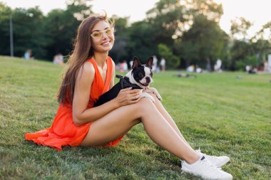 Mutlu güzel bir kadın yaz parkında çimenlerde oturuyor, elinde Boston Terrier köpeği, gülümsüyor pozitif ruh hali, turuncu elbise giyiyor, moda tarzı, ince bacaklar, spor ayakkabıları, evcil hayvanlarla oynuyor.