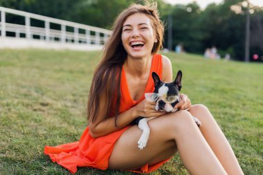 Yaz parkında çimenlerde oturan bir kadın elinde Boston Teriyeri, komik bir köpek şık güneş gözlüğü takan bir kız evcil hayvanla oynuyor, eğleniyor, gülüyor, pozitif ruh hali içinde.