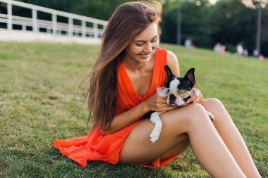 Yaz parkında çimenlerde oturan, Boston Terrier köpeği tutan, gülümseyen, turuncu elbise giyen, moda tarzı olan, güneş gözlüklü mutlu bir kadının portresi.