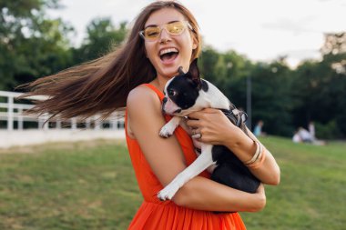 Genç ve mutlu bir kadın Boston terrier köpeğini parkta tutuyor, yaz güneşli bir gün, neşeli bir ruh hali, evcil hayvanla oynamak, uzun saçlar sallamak, eğlenmek, güneş gözlüğü takmak, gülmek.