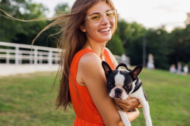 Turuncu elbiseli, gülümseyen genç kadın parkta köpekle oynarken eğleniyor, yaz stili, neşeli bir ruh hali.