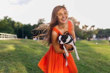 Boston Terrier köpeği tutan mutlu güzel kadın parkı, gülümseyen pozitif ruh hali, modaya uygun yaz tarzı, turuncu elbise, güneş gözlüğü takan, evcil hayvanla oynayan, eğlenen, güneşli hafta sonu eğlencesi