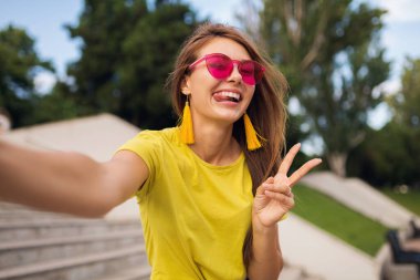 Şehir parkında fotoğraf çeken genç, şık, güzel bir kadın. Pozitif, duygusal, sarı bluz, pembe güneş gözlüğü takan, yaz tarzı moda eğilimi olan, uzun saçlı, barış işareti gösteren.