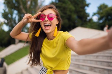 Şehir parkında fotoğraf çeken genç, şık, güzel bir kadın. Pozitif, duygusal, sarı bluz, pembe güneş gözlüğü takan, yaz tarzı moda eğilimi olan, uzun saçlı, barış işareti gösteren.