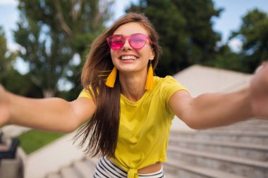 Şehir parkında fotoğraf çeken genç, şık, güzel bir kadın. Pozitif, duygusal, sarı bluz, pembe güneş gözlüğü takan, yaz tarzı moda eğilimi olan, uzun saçlı, eğlenen.