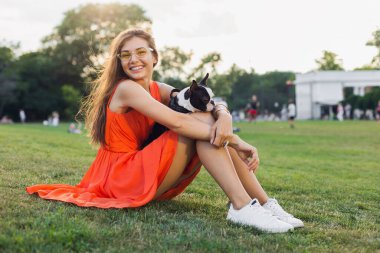 Mutlu güzel bir kadın yaz parkında çimenlerde oturuyor, elinde Boston Terrier köpeği, gülümsüyor pozitif ruh hali, turuncu elbise giyiyor, moda tarzı, ince bacaklar, spor ayakkabıları, evcil hayvanlarla oynuyor.