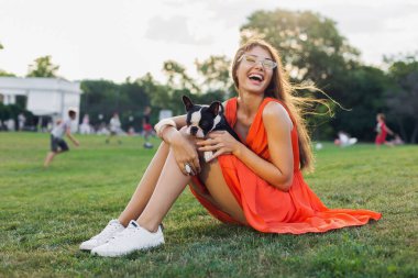 Mutlu gülen güzel bir kadın yaz parkında çimenlerde oturuyor, Boston Terrier köpeği tutuyor, gülümsüyor pozitif ruh hali içinde, turuncu elbise giyiyor, moda tarzı, ince bacaklar, evcil hayvan ayakkabılarıyla oynuyor.