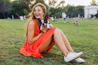 Yaz parkında çimenlerde oturan, Boston Terrier köpeği tutan, gülümseyen, turuncu elbise giyen, moda tarzı, ince bacakları olan, evcil hayvanla oynayan güzel bir kadın.
