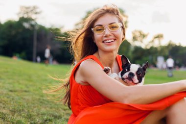 Turuncu elbiseli, gülümseyen genç kadın parkta köpekle oynarken eğleniyor, yaz stili, neşeli bir ruh hali.