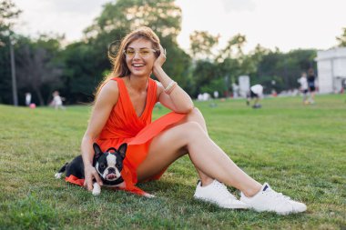 Mutlu güzel bir kadın yaz parkında çimenlerde oturuyor, elinde Boston Terrier köpeği, gülümsüyor pozitif ruh hali, turuncu elbise giyiyor, moda tarzı, ince bacaklar, spor ayakkabıları, evcil hayvanlarla oynuyor.