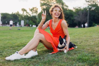 yaz parkında çimenlerde oturan pozitif kadın, Boston Terrier köpeği tutuyor, komik surat ifadesi, turuncu elbise giyiyor, moda tarzı, ince bacaklar, spor ayakkabıları, evcil hayvanlarla oynuyor.