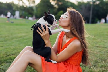 Yaz parkında çimenlerde oturan, Boston Terrier köpeği tutan, gülümseyen, turuncu elbise giyen, moda tarzı olan, güneş gözlüklü mutlu bir kadının portresi.