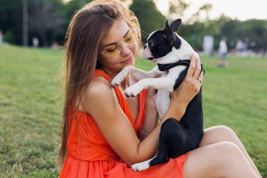 Yaz parkında çimenlerde oturan, Boston Terrier köpeği tutan, gülümseyen, turuncu elbise giyen, moda tarzı olan, güneş gözlüklü mutlu bir kadının portresi.