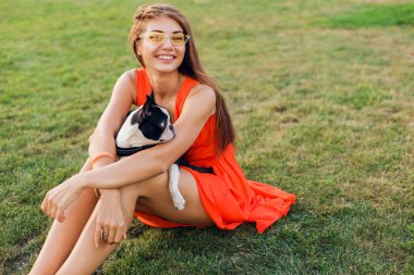 Turuncu elbiseli, gülümseyen genç kadın parkta köpekle oynarken eğleniyor, yaz stili, neşeli bir ruh hali.