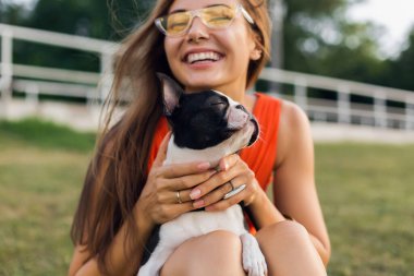 Yaz parkında çimenlerde oturan, Boston Terrier köpeği tutan, gülümseyen, turuncu elbise giyen, moda tarzı olan, güneş gözlüklü mutlu bir kadının portresi.