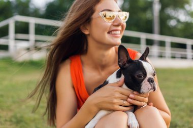Turuncu elbiseli, gülümseyen genç kadın parkta köpekle oynarken eğleniyor, yaz stili, neşeli bir ruh hali.
