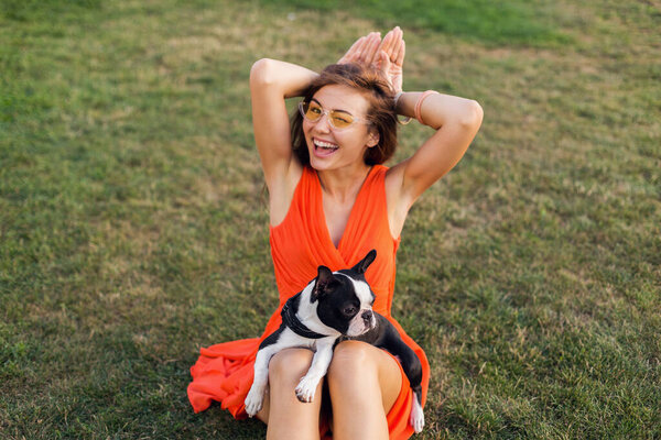 portrait of happy pretty woman sitting on grass in summer park, holding boston terrier dog, smiling positive mood, wearing orange dress, trendy style, sunglasses, playing with pet