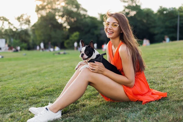 幸せな美しい女性は夏の公園で草の上に座って ボストンのテリア犬を保持し 肯定的な気分笑顔 オレンジのドレスを着て 流行のスタイル スリムな足 スニーカー ペットと遊ぶ — ストック写真
