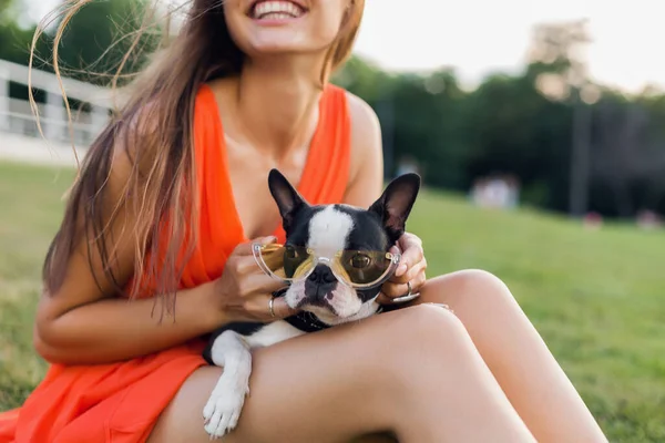 夏の公園で草の上に座って幸せな女性の肖像画 ボストンのテリア犬を保持し 肯定的な気分笑みを浮かべて オレンジのドレス 流行のスタイル サングラスを着て ペットと遊ぶ — ストック写真