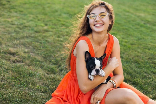 公園で草の上に座って魅力的な若い女性 夏のスタイル オレンジのドレスを身に着けています ボストンテリア犬を保持します 積極的 スタイリッシュなサングラス — ストック写真