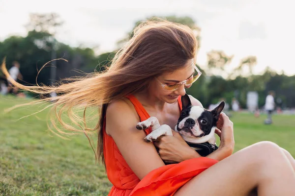 夏の公園で草の上に座って幸せな女性の肖像画 ボストンのテリア犬を保持し 肯定的な気分笑みを浮かべて オレンジのドレス 流行のスタイル サングラスを着て ペットと遊ぶ — ストック写真