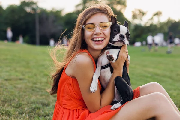 夏の公園で草の上に座って幸せな女性の肖像画 ボストンのテリア犬を保持し 肯定的な気分笑みを浮かべて オレンジのドレス 流行のスタイル サングラスを着て ペットと遊ぶ — ストック写真