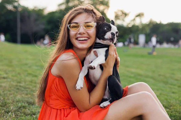 夏の公園で草の上に座って幸せな女性の肖像画 ボストンのテリア犬を保持し 肯定的な気分笑みを浮かべて オレンジのドレス 流行のスタイル サングラスを着て ペットと遊ぶ — ストック写真