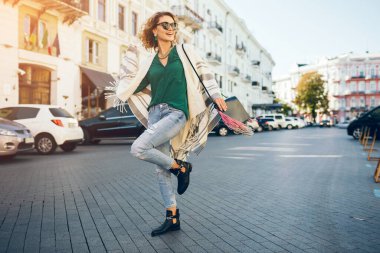 Havalı pozitif kadın baharda sokakta yürüyor, moda trendi, kot pantolon, pelerin, yeşil bluz, şık aksesuarlar, mutlu duygu