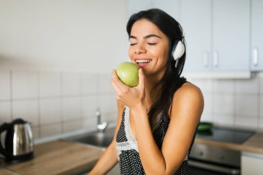 Sabah mutfakta yemek pişiren, elma yiyen, gülümseyen, mutlu, pozitif bir ev kadını sağlıklı bir yaşam tarzı olan, kulaklıkla müzik dinleyen, gülen, beyaz dişli genç bir kadının portresi.