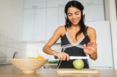 Genç ve çekici bir kadın sabah mutfakta yemek pişiriyor, meyve salatası yapıyor, elmayı bıçakla kesiyor, gülümsüyor, mutlu bir ruh hali içinde, pozitif bir ev hanımı, sağlıklı bir yaşam tarzı, kulaklıkla müzik dinliyor.,
