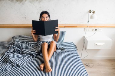 Sabahları yatakta oturmuş kulaklıkla müzik dinleyen ve kitap okuyan çok seksi bir kadın.
