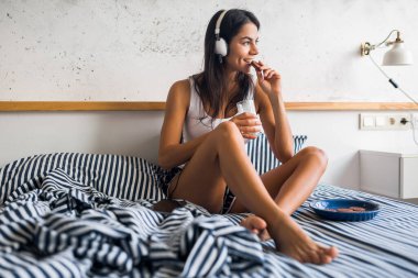 Genç gülümseyen esmer kadın, sabahları yatakta oturup kurabiye yiyor, süt içiyor, kulaklıkla müzik dinliyor, mutlu ruh hali, uyanıyor, rahat bir şekilde pijamalarını giyiyor, rahatlıyor, rahatlıyor, kahvaltı ediyor.