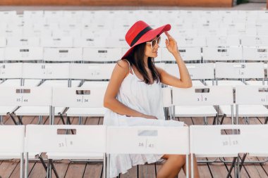 Beyaz elbiseli, kırmızı şapkalı, güneş gözlüklü çekici, mutlu bir kadın. Yazın açık hava tiyatrosunda tek başına oturuyor. Spring Street tarzı moda modası.