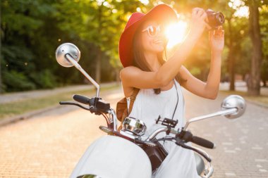 Sokakta motosiklet süren çekici bir kadın, yaz tatili tarzı, seyahat eden, gülümseyen, mutlu, eğlenen, şık kıyafetler, maceralar, klasik fotoğraf makinesinde fotoğraf çeken.