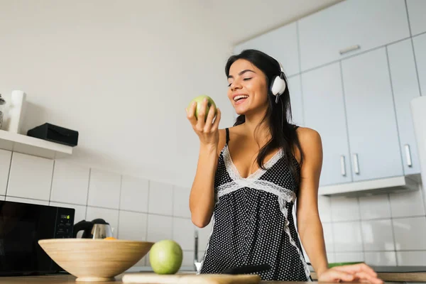 attractive smiling woman in sexy pajamas having breakfast in kitchen in morning, healthy lifestyle, eating apple, listening to music on headphones