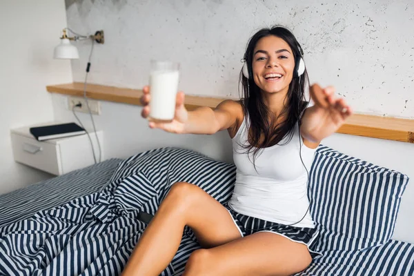 attractive smiling skinny woman in pajamas lying in bed at home having rest wake up in morning listening to music on headphones having breakfast eating cookies and drinking milk
