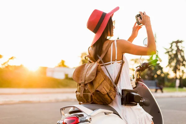 attractive smiling woman riding on motorbike in street in summer style outfit wearing white dress and red hat traveling on vacation, taking pictures on vintage photo camera