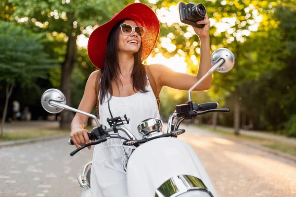 attractive smiling woman riding on motorbike in street in summer style outfit wearing white dress and red hat traveling on vacation, taking pictures on vintage photo camera