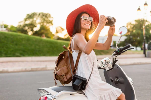 attractive smiling woman riding on motorbike in street in summer style outfit wearing white dress and red hat traveling on vacation, taking pictures on vintage photo camera