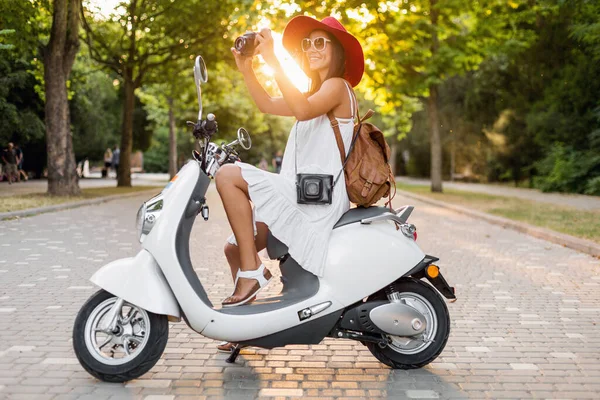 白いドレスを着た夏のスタイルの服装でバイクに乗っている魅力的な笑顔の女性と休暇中に旅行する赤い帽子 ヴィンテージフォトカメラで写真を撮る — ストック写真