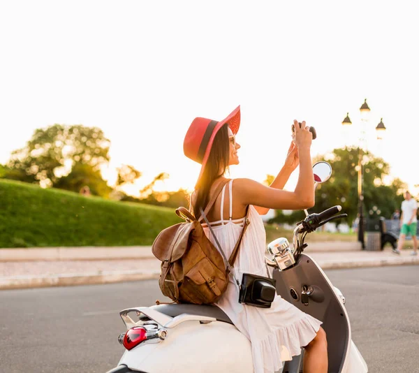 白いドレスを着た夏のスタイルの服装でバイクに乗っている魅力的な笑顔の女性と休暇中にバックパックで旅行する赤い帽子 ヴィンテージフォトカメラで写真を撮る — ストック写真