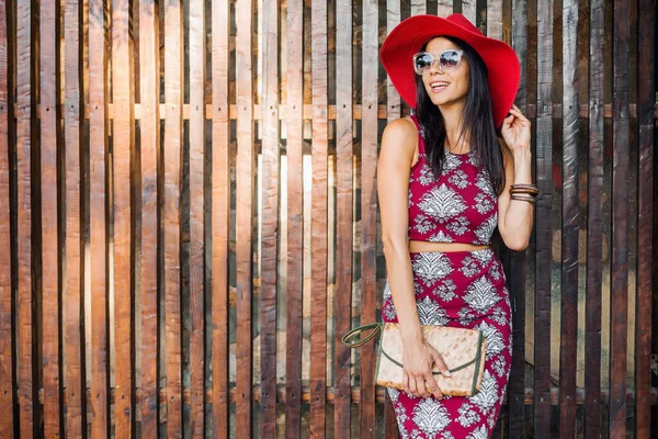 夏のファッショントレンド トップ スカート スキニー わらハンドバッグ 赤い帽子 サングラス アクセサリー 幸せな気分 休暇で木製の壁に対してポーズスタイリッシュな美しい女性 — ストック写真
