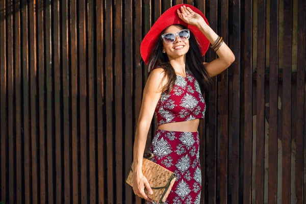 スタイリッシュな美しい女性は 印刷された服で木製の背景にポーズ 夏のスタイルのファッショントレンド トップ スカート スキニー わらハンドバッグ 赤い帽子 サングラス アクセサリー — ストック写真