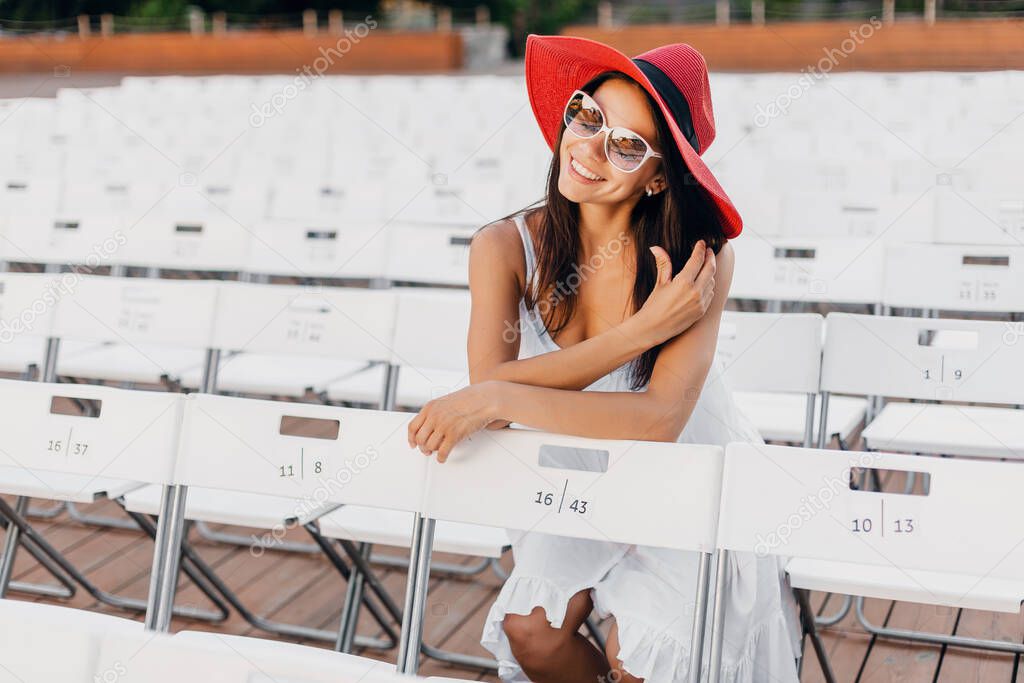 attractive woman dressed in white dress, red hat, sunglasses sitting in summer open air theatre on chair alone, spring street style fashion trend