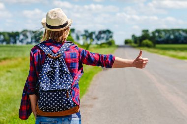 Şapkalı ve sırt çantalı bir kız yolda otostop çekiyor.