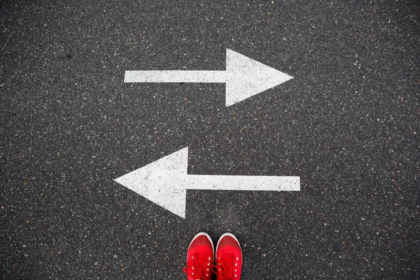 2 つの方向に描かれた矢印を持つアスファルトの道路に赤いスニーカー。意思決定や選択肢を作る — ストック写真