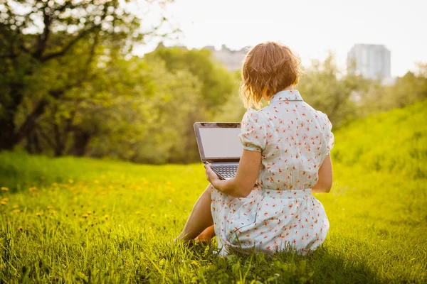 Giovane donna con computer portatile seduta su erba verde nel parco al tramonto — Foto Stock