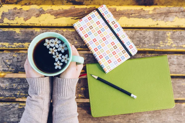 Mujer sosteniendo taza caliente de té de primavera con cuaderno y lápiz sobre fondo de madera — Foto de Stock