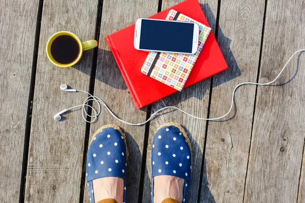 Hora de trabajar / estudiar. Estilo de vida, vacaciones de verano, educación y concepto de personas. Pies de niña hipster joven con taza de café, teléfono inteligente, auriculares, libros apilados sobre fondo de madera — Foto de Stock